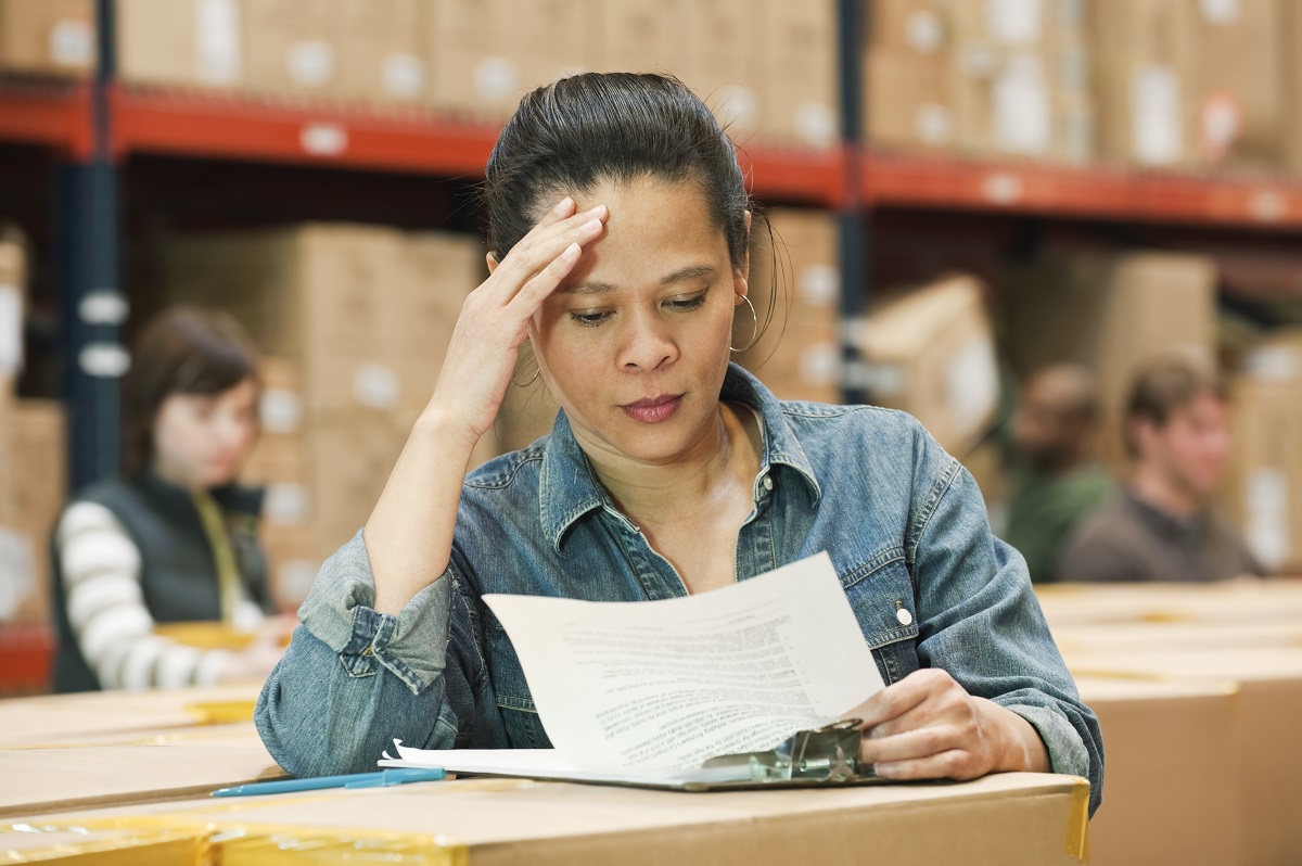 African American female warehouse worker working on inentory on a lap top computer in a large distribution warehouse of products stored in cardboard boxes and on large racks.